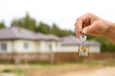 Anahtarlı bir el ve ahşap bir anahtarlık. Çitlerin ve kulübenin arka planı. Bina, proje, yeni bir eve taşınma, ipotek, kira ve emlak satın alma. Kapıyı açmak için. Boşluğu kopyala