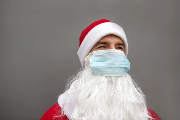 Santa Claus with white beard in red coat in a medical mask. Christmas in the coronavirus pandemic, seasonal diseases, SARS and pneumonia in the holidays. Protection from viruses in the new year. Gray background