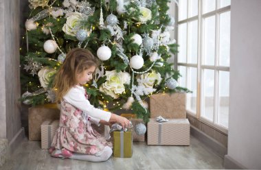 Güzel elbiseli küçük kız elinde hediye kutusuyla Noel ağacının altında oturuyor. Büyük bir pencereden ışık, karanlık bir oda, ev konforu, Noel, sihir ve neşe. Yeni yıl, beyaz dekor