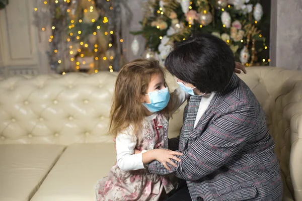 Grandmother and granddaughter on the sofa in the living room with Christmas decor hugging in medical masks on their faces. A family holiday during the outbreak of coronavirus and disease. New Year