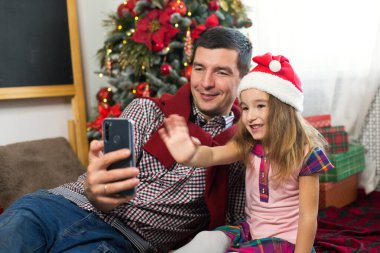 Baba ve kızı akıllı telefonlu Noel ağacının yanında selfie çekiyorlar, video bağlantısıyla iletişim kuruyorlar. Noel kutlaması, hediye kutusu, Noel Baba şapkalı bir kız el sallıyor. Yeni yıl, tatil dekoru