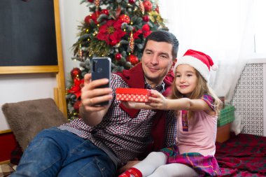 Baba ve kızı akıllı telefonlu Noel ağacının yanında selfie çekiyorlar, video bağlantısıyla iletişim kuruyorlar. Noel kutlaması, hediye kutusu, Noel Baba şapkalı bir kız el sallıyor. Yeni yıl, tatil dekoru