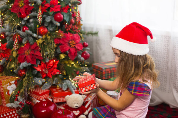 Little girl in Santa hat opens a red box with a gift and a Golden magic light near the Christmas tree. Holiday decor, poinsettias on fir trees, New year. Joy, surprise, children's emotions. Copyspace