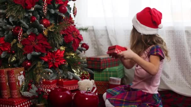 戴着圣诞礼帽的小女孩在圣诞树旁打开一个装有礼物和金色魔法灯的红色盒子 假日装饰 杉树上的一品红 孩子们的情绪 — 图库视频影像