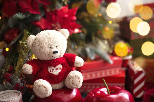 White Teddy bear in red knitted sweater with heart on the chest and the words Love near Christmas tree among the gift boxes. Gift to your beloved, Declaration of love, poinsettia decor. Copy space