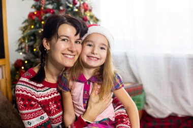 Anne ve kız kucaklaşır, mutlu bir şekilde gülümser ve kameraya bakar, Noel ağacının yanında rahat giysiler ve Noel Baba şapkasıyla otururlar. Noel, evde yeni yıl arifesi. Dekor, hediye kutuları, kopyalama alanı