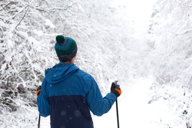 Rüzgârlık giymiş kayakçı ve elinde ponponlu, sırtında kayak sopalarıyla karlı bir ormanın arka planına yaslanmış bir şapka. Kros kayağı kış ormanlarında, açık hava sporlarında, sağlıklı yaşam tarzında..