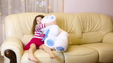 Happy girl in a pink dress hugging a big white soft bear home on the couch. Playing with lollipops heart. Gift from a Mother, valentine's day, birthday, holiday. Copy space