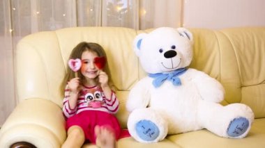 Happy girl in a pink dress hugging a big white soft bear home on the couch. Playing with lollipops heart. Gift from a Mother, valentine's day, birthday, holiday. Copy space