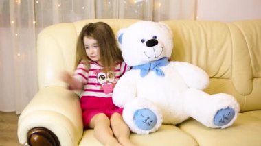 Happy girl in a pink dress hugging a big white soft bear home on the couch. Playing with lollipops heart. Gift from a Mother, valentine's day, birthday, holiday. Copy space
