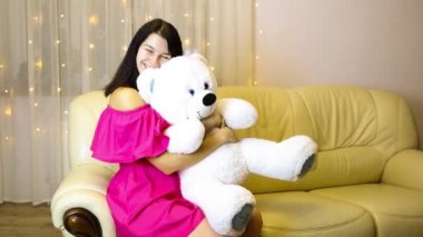 Happy girl in a pink dress hugging a big white soft bear home on the couch. Gift from a loved one, valentine's day, birthday, holiday. Copy space