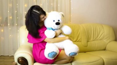 Happy girl in a pink dress hugging a big white soft bear home on the couch. Gift from a loved one, valentine's day, birthday, holiday. Copy space