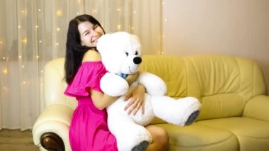 Happy girl in a pink dress hugging a big white soft bear home on the couch. Gift from a loved one, valentine's day, birthday, holiday. Copy space