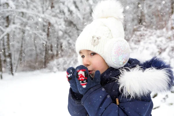 A little girl in the cold warms her hands in mittens-breathes warm air from her mouth on her hands. Winter, walk the baby in the open air, the snow. Warm clothing, knitted hat, fur pompom and hood