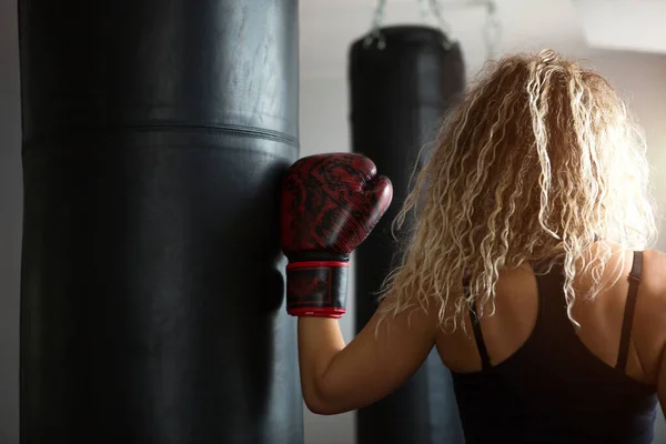 パンチングバッグの近くにボクシンググローブを持つ長い巻きブロンドの髪を持つ女性 ボクシングに従事 健康的なライフスタイル 運動体 持久力 スペースのコピー — ストック写真