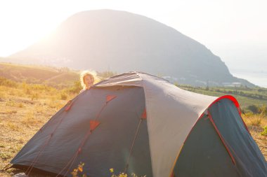 Neşeli çocuk şafak vakti panoramik manzaralı dağlardaki bir turist çadırının arkasından dışarı bakar. Ev turizmi, aktif yaz gezisi, aile maceraları. Ekoloji, kamp, dağ yürüyüşü..