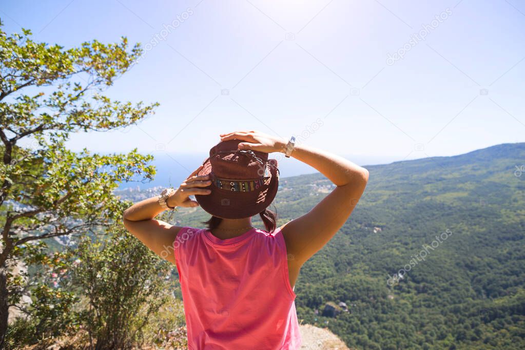 Woman in a hat looks at the panoramic view from the mountain to the sea and the forest. Tourist, trekking, travel. Active ecotourism, healthy lifestyle, adventure