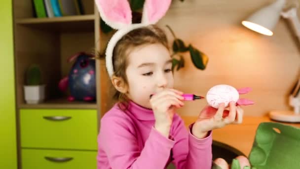 頭にウサギの耳を持つ女の子は テーブルの上にフェルトのペンで白い卵を描きます イースターの準備 子供のための教育活動 子供に興味を持つ方法 イースターバニー Diyクラフト — ストック動画