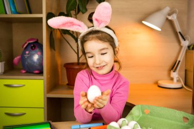 Paskalya tavşanının kulaklarında bir kız ev içinde keçeli kalemle yumurta boyuyor. El işleri, dini bayram hazırlıkları, yumurtalı bir tepsi, plastik kulaklı tavşan.