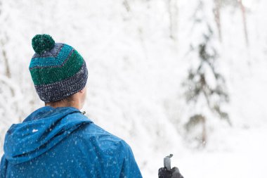 Rüzgârlık giymiş kayakçı ve elinde ponponlu, sırtında kayak sopalarıyla karlı bir ormanın arka planına yaslanmış bir şapka. Kros kayağı kış ormanlarında, açık hava sporlarında, sağlıklı yaşam tarzında..