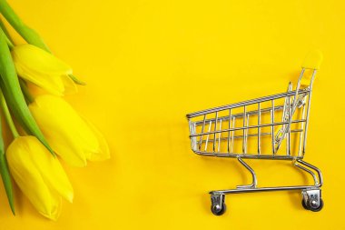 Katı renk arka planda sarı laleler ve alışveriş arabası. Bahar ve yaz çevrimiçi alışveriş, bayram, Paskalya, kadınlar günü, 8 Mart, çiçekçi, çiçekçi. Boşluğu kopyala