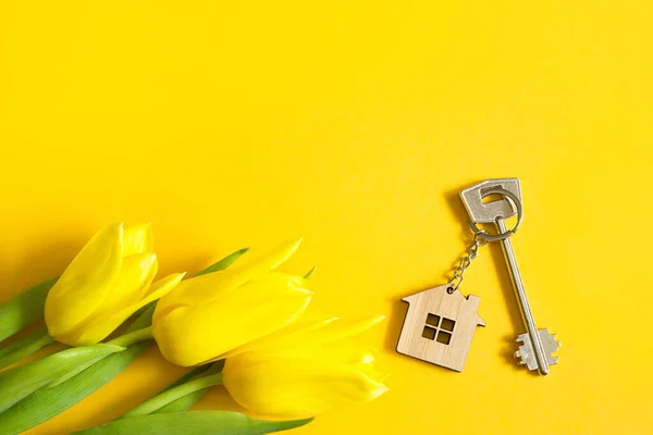 Key ring in the shape of wooden house with key on yellow background and spring tulips. Building, design, project, moving to new home, mortgage, rent and purchase real estate, summer offer. Copy space