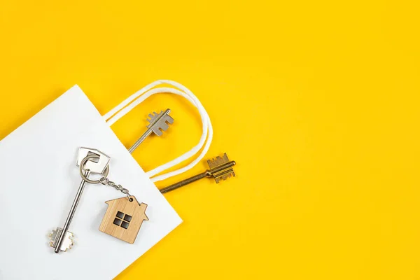 Key ring in the shape of wooden house with key on a yellow background in a white paper gift wrapping bag. Mortgage, rent and purchase real estate, beneficial offer for construction. Copy space