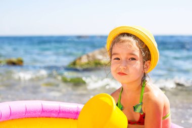 Sarı hasır şapkalı kız deniz kenarında şişme bir havuzda oturuyor ciddi ve tatminsiz bir bakışla. Çocukların cildini güneşten korumak için, güneş yanığı. Deniz kıyısında tatil yeri.