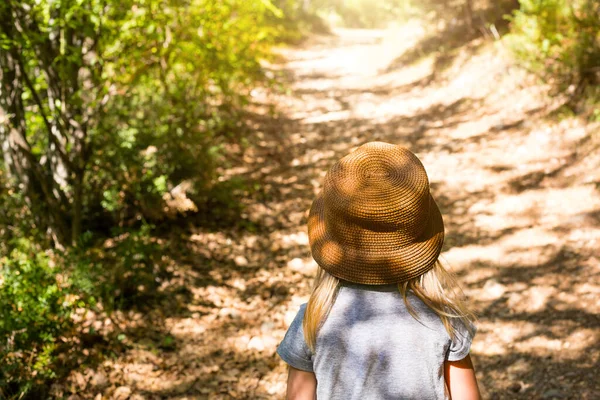 A little girl in a straw hat walks along a path in the forest with sunlight and shadows from the trees. View from the back, the road ahead. A tourist path in the fresh air. Lonely child lost, danger