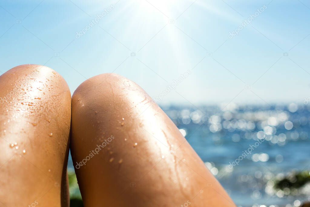 Smooth tanned female legs in the spray of water on the beach against the background of the sea. Tanning products, sunburn protection, skin care, depilation, beach holidays. Copy space