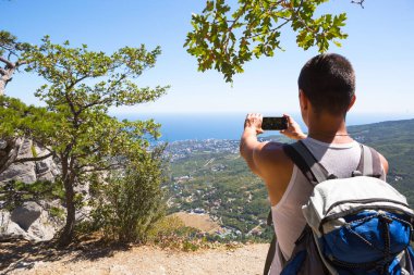 Erkek turist, en tepeden kozalaklı ağaçların arasına dağlık bir alanda deniz manzarası olan akıllı bir telefondan fotoğraf çekiyor. Video iletişimi, seyahat ederken telefonu kullanmak. Gezintide SIM kart