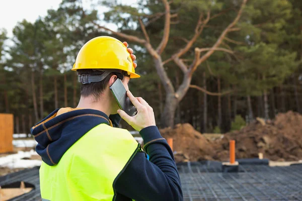 İnşaat işçisi, vakıf, borular ve kör alan inşaatının arka planına karşı sarı kasklı ve koruyucu yelekli akıllı bir telefonla konuşuyor..