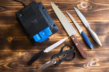 Evdeki elektrikli bileyiciye bıçak keskinleştirmek. Tahta bir masanın üzerinde bileme makinesi olan bıçak ve makas düzeni. Üst manzara, düz uzanma. 