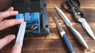 Evdeki elektrikli bileyiciye bıçak keskinleştirmek. Adamın eli bıçak bıçağını mavi bileyicilerin arasından geçirir, makinede toz bulutu uçuşur. Kaluga, Rusya, 25 Nisan 2021