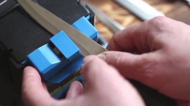 Evdeki elektrikli bileyiciye bıçak keskinleştirmek. Adamın eli bıçak bıçağını mavi bileyicilerin arasından geçirir, makinede toz bulutu uçuşur. Kaluga, Rusya, 25 Nisan 2021