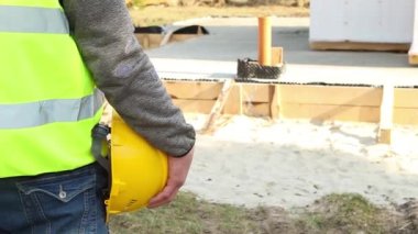 İnşaatçının eli inşaat sahasında ahşap işçilik ve inşaat malzemeleriyle evin temelini oluşturan sarı koruyucu bir şapka tutuyor. İş güvenliği.