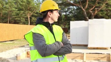 Koruyucu sarı kasklı ve işaret yeleği giyen bir inşaat işçisi, temeli ve havalandırmalı beton blokları olan bir evin inşaat alanının önünde duruyor. Foreman, inşaat malzemeleri..