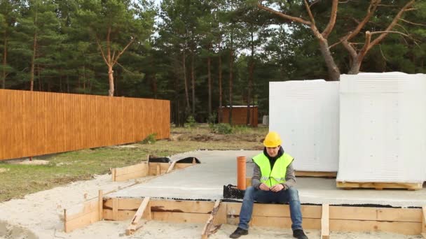 保護された黄色の硬い帽子と信号ベストの建設労働者は 木製の型枠と建設現場のコンクリートブロックを通気して基礎に座っています フォアマン建築材料 — ストック動画