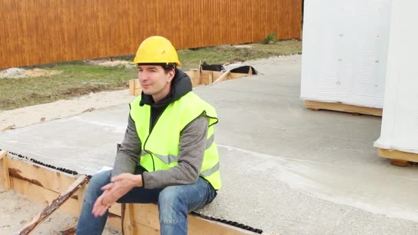 保護された黄色の硬い帽子と信号ベストの建設労働者は 木製の型枠と建設現場のコンクリートブロックを通気して基礎に座っています フォアマン建築材料 — ストック動画