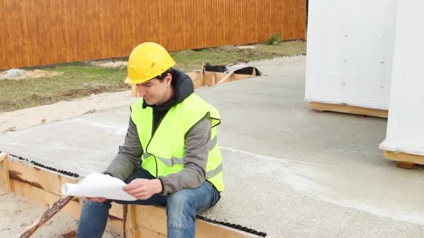 身穿黄色防护帽和信号弹背心的建筑工人在建筑工地用木制模板在地基上坐下来 研究图纸和文件 建筑材料 — 图库视频影像
