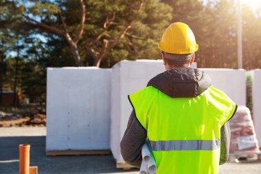 Proje mimarının sırtı, temelleri atılmış ve bloklar teslim edilmiş bir evin inşaat sahasında. Sarı baret ve işaret yelekli inşaat işçisi. Model 