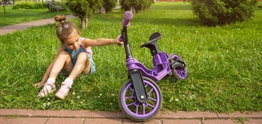 Yazın mor koşu bisikletinin yanındaki parkta çimlerin üzerinde dinlenen neşeli kız. Çocukların aktif eğlencesi, yürümeye başlayan çocuklar için scooter, mutlu çocuk. Uzayı kopyala Yaz arkaplanı