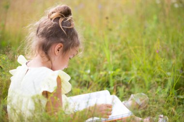 Sarı elbiseli bir kız tarladaki bir battaniyenin üzerinde çimenlerde oturur ve bir kağıt kitap okur. Uluslararası Çocuk Günü. Yaz zamanı, çocukluk, eğitim ve eğlence, kır evi özü. Boşluğu kopyala