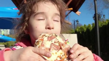 Kız komik ve açık hava parkındaki bir kafede iştahlı pizza yiyor. Sokak yemeği, aile zamanı, yürüyüş.