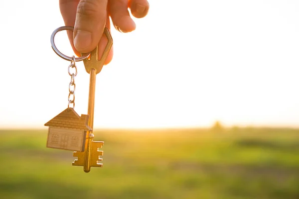 House key with keychain in hand. Background of sky, sunlight and field. Dream of home, building a cottage in countryside, plan and project design, farm, moving to a new home, accommodation. Copy space