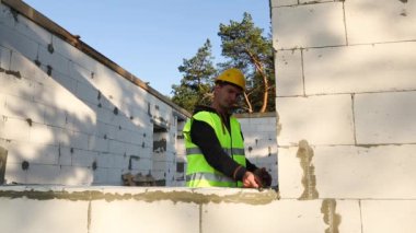 İnşaat işçisi, pencere açıklığının uzunluğunu ve duvarın uzunluğunu mezurayla ölçüyor. Kulübeler havalandırılmış beton bloklar, koruyucu giysiler - kalın şapka ve bir yelek