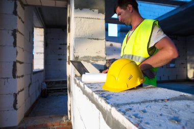 Gözenekli beton bloklardan yapılmış bir evin şantiyesinde inşaat mühendisi yansıtıcı güvenlik yeleği ve baret takan bir bilgisayarda çalışıyor. Tasarım, inşaat, çizim projesi, kontrol