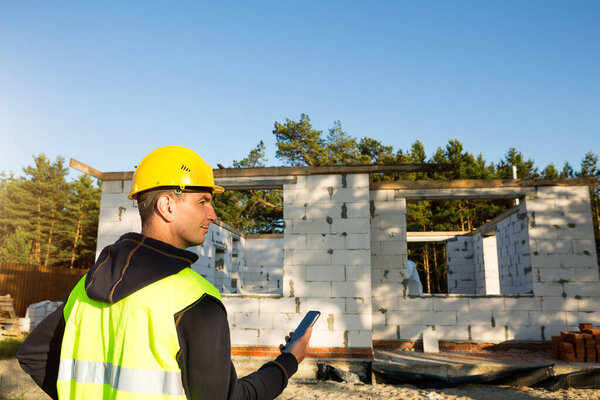 Construction worker talks on a smartphone in a yellow hardhat and reflective vest against the background of the construction of house-the walls and window openings are made of porous concrete blocks.
