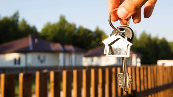 Key ring with keychain house in hand. Background of fence and cottage. Moving to a new home, mortgage, buying real estate, renting and booking housing, dream of living  in the village. Copy space