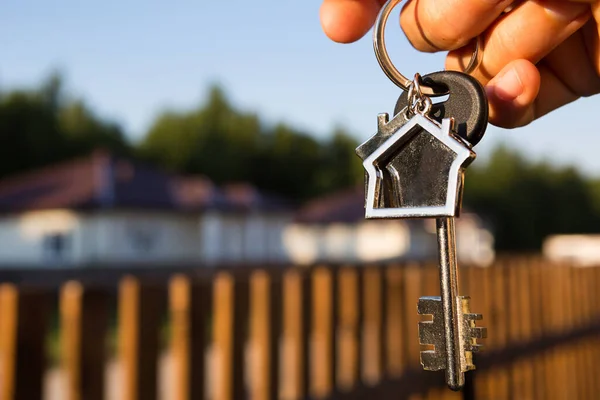 Key ring with keychain house in hand. Background of fence and cottage. Moving to a new home, mortgage, buying real estate, renting and booking housing, dream of living  in the village. Copy space